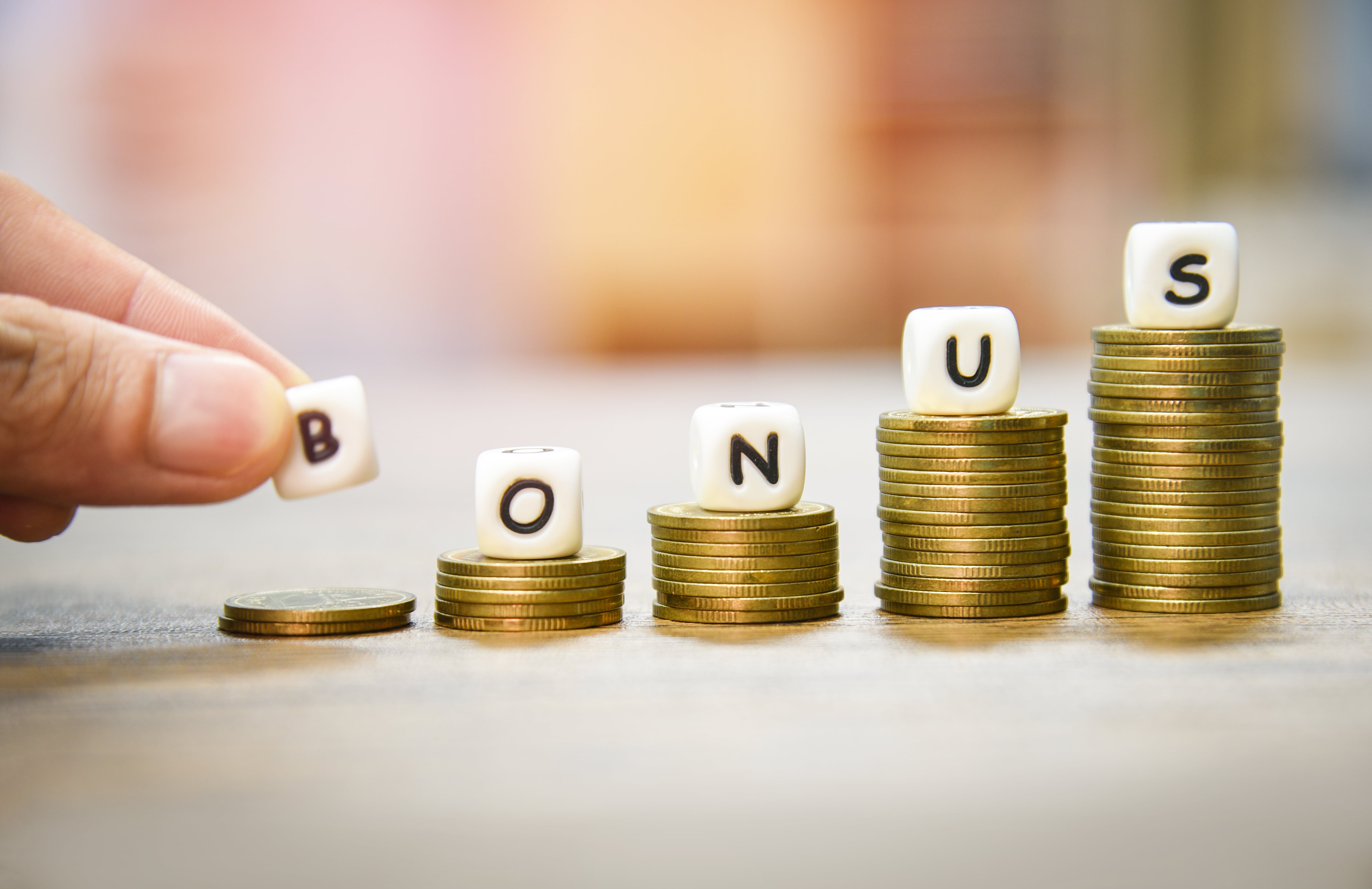 five small white dice spelling the word 'BONUS' are laid on top of stacks of gold coins that increase in size from left to right, the 'B' on the shortest, leftmost stack is still being placed by a person's hand. 