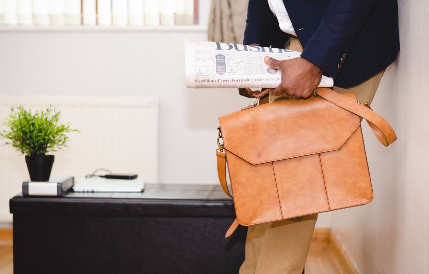 An industrious entrepreneur or business owner leaning against an office wall with a business newspaper in hand along with a tan briefcase.
