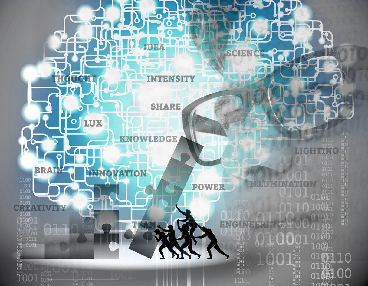 An image with several layers: the face of a confident entrepreneur examining a variety of ideas expressed as terms, while puzzle pieces are placed together by a team of individuals, a network of lights which resemble the human brain flicker, and, in the very back of the image, 1s and 0s are displayed in various sizes and combinations.