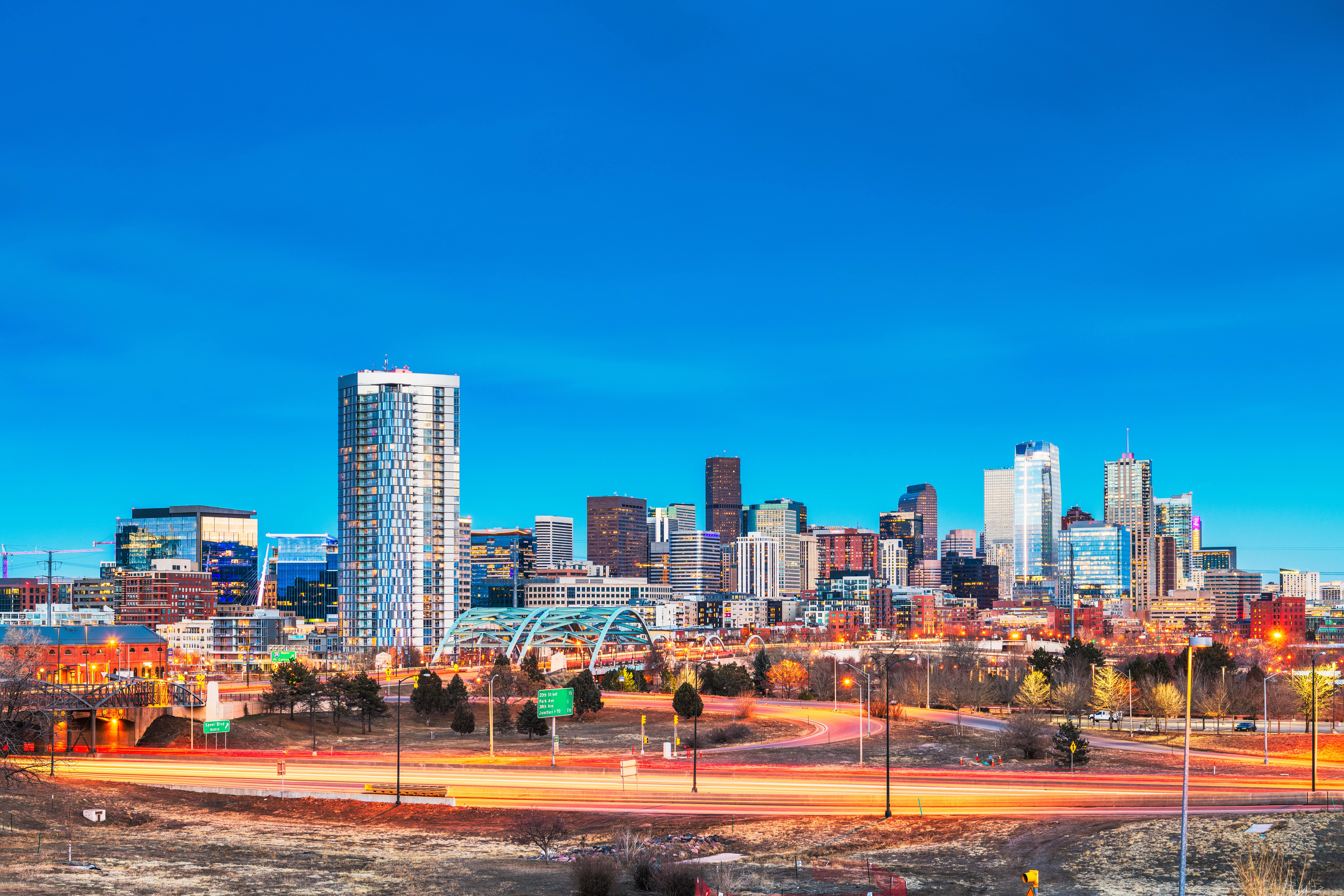 a time lapse view of downtown Denver, Colorado at dusk with the city lit up in the background under a dark blue sky with the lights of the highway blurred out in front. 