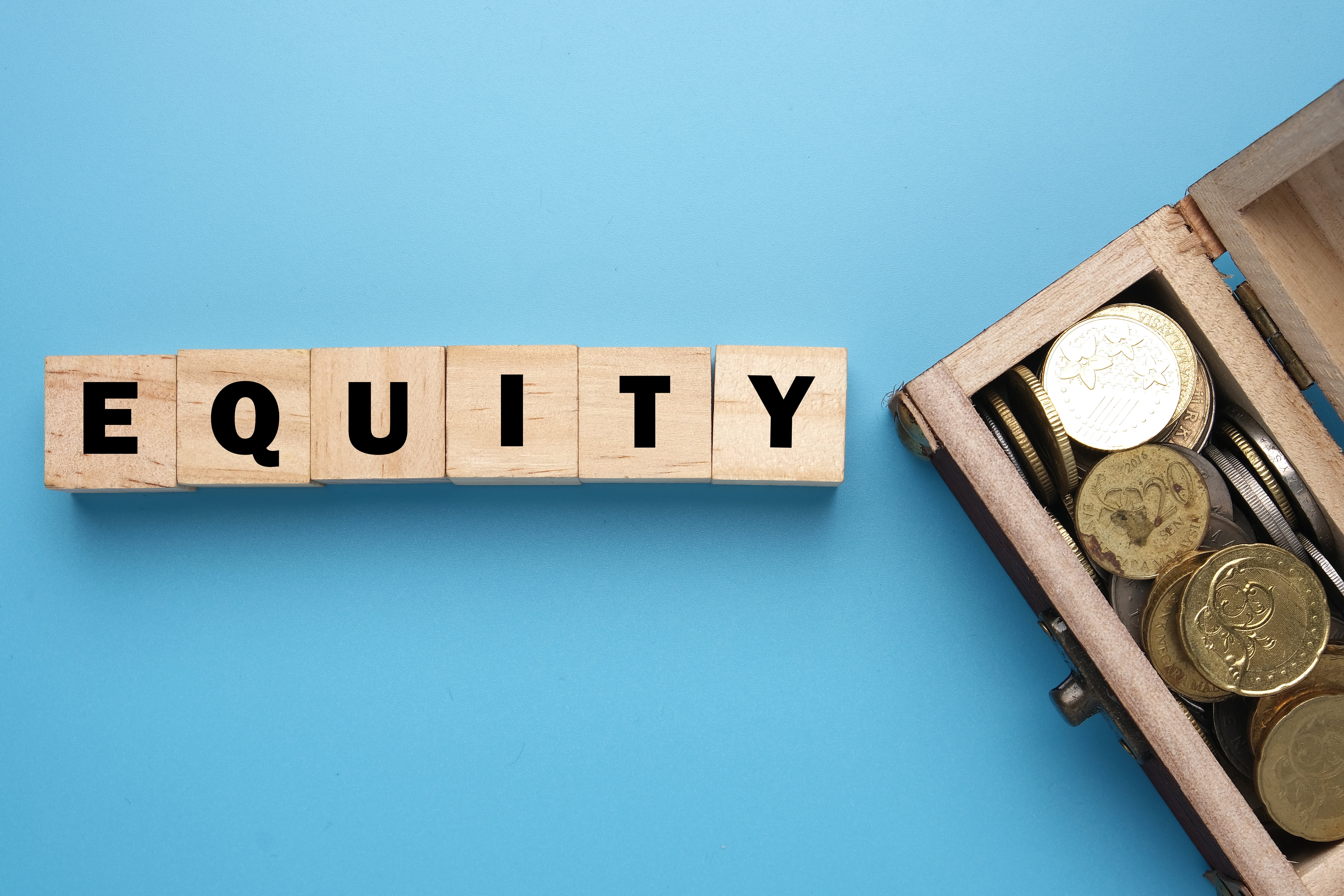 The words 'EQUITY' appears in wooden block letters with black text next to a wooden box full of coins, and both on top of a sky blue background.
