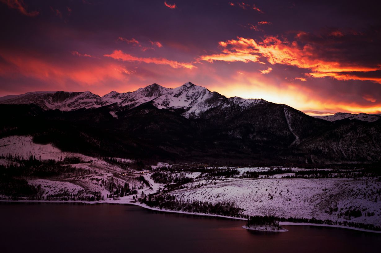 The sky is ablaze at sunset over the snowy Rocky Mountains in Colorado, towering above a still Alpine lake.