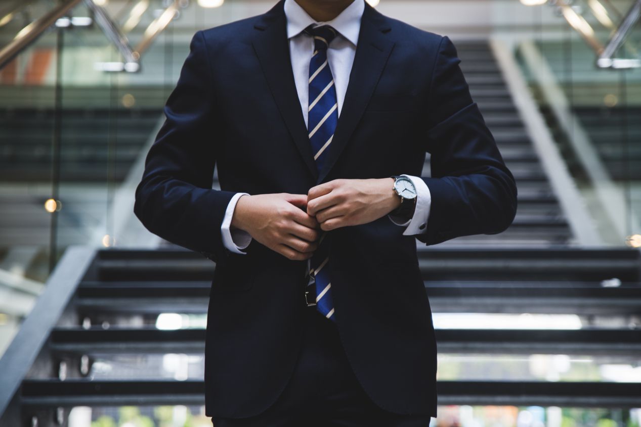 Close up of a businessman's torso - he's wearing a navy suit and tie and stands before a glass staircase.