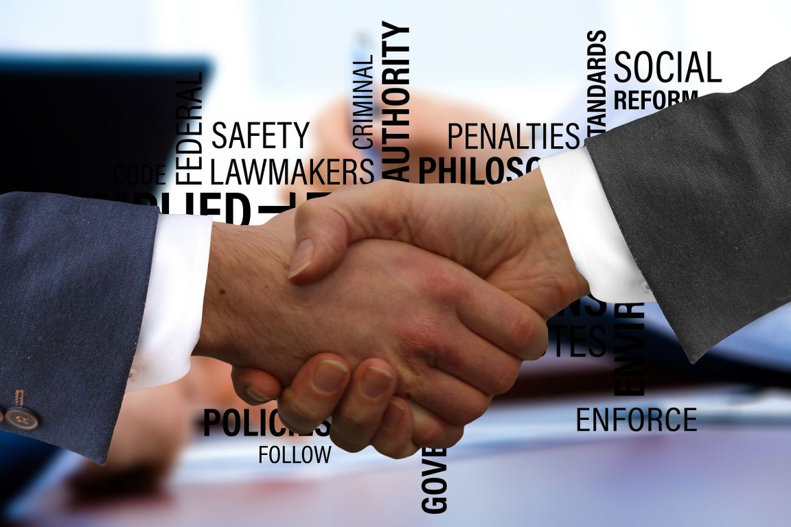 A firm handshake between two business people in front of important terms - like policies, standards, governance, and lawmakers - all arranged variously and in a variety of sizes.